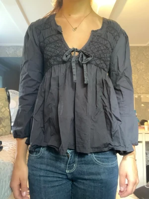 Svart blus med smock och rosett - Snygg svart blus med smock upptill och en söt rosettdetalj vid urringningen. Blusen har lång ärm med lätt puff och avslappnad passform. Tillverkad i ett tunt, mjukt tyg som ger ett flowy intryck. 