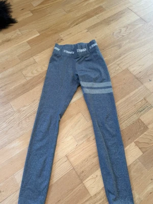 Gråa träningsbyxor från Stronger. - Snygga grå leggings från Stronger med bred resår i midjan där loggan STRONGER syns runtom. På ena benet finns två vita randiga detaljer med prickigt mönster. Stretchigt material som sitter bekvämt på. 