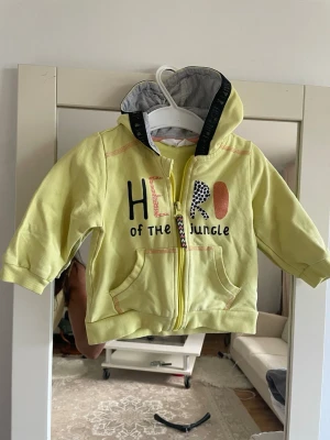 Gul hoodie med tryck och huva - Snygg gul hoodie för barn med texten 'HERO of the JUNGLE' framtill och 'SUPER' på ryggen. Hoodien har dragkedja, två fickor fram och en gråmelerad insida i huvan. Perfekt för dig som gillar färg och coola prints.