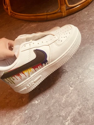 Vita sneakers med färgstänk - Unika vita sneakers med svart swoosh och färgglada droppdetaljer i rött, gult, grönt och blått på sidan. Klassisk låg modell med perforerad tå och vit sula. Skorna är i syntetmaterial och kommer med extra vita och grå skosnören.