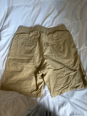 Beige shorts med snörning från H&M - Säljer ett par beige shorts från H&M med elastisk midja och snörning framtill. Shortsen har två bakfickor och är tillverkade i ett mjukt bomullsmaterial. Perfekta för varma dagar och enkel att matcha med olika stilar.
