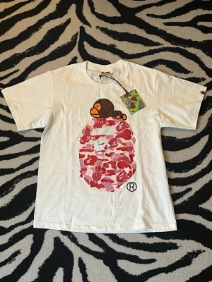 Vit BAPE t-shirt med camomönster - Vit t-shirt från BAPE med stort tryck av det klassiska apansikte-motivet i rosa och rött camomönster på bröstet. Ovanpå motivet ligger en liten apa. T-shirten har korta ärmar och rund hals. Perfekt för dig som gillar streetwear och unika prints.