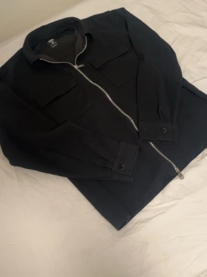 Svart overshirt med dragkedja FSBN - Snygg svart overshirt från FSBN i storlek XS. Overshirten har två bröstfickor med lock, silverfärgad dragkedja framtill och klassisk krage. Tillverkad i ett kraftigare tyg som ger en stilren look. Perfekt att bära över en t-shirt eller hoodie.