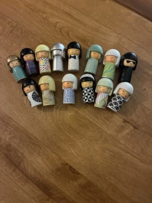 Samling små kokeshi-dockor - Säljer en samling med 16 små kokeshi-dockor i olika färger och mönster. Dockorna är gjorda av trä och plast, och har unika frisyrer och kläder i allt från pastell till svartvitt och metallic. Perfekt för dig som gillar japansk design eller vill ha fina dekorationer.