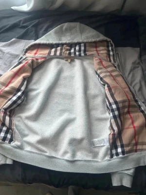 Burberry hoddie  - Säljer en grå hoodie med dragkedja och huva. Insidan är fodrad med ett klassiskt beige, svart och rött rutmönster. Hoodien har ribbade muddar och känns riktigt mjuk och skön. Perfekt för dig som gillar stilrena detaljer och vill ha något som sticker ut lite extra.