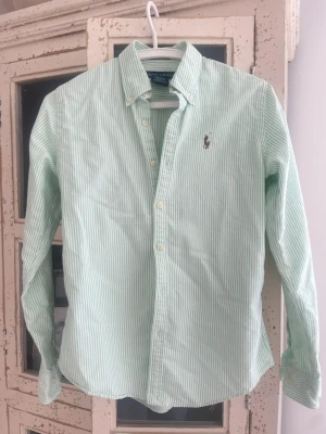 Grön- och vitrandig skjorta Ralph Lauren - Snygg grön- och vitrandig skjorta från Ralph Lauren med klassisk krage och knappar framtill. Skjortan har lång ärm och den ikoniska broderade loggan på bröstet. Perfekt för en clean och stilren look.