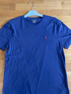 Blå t-shirt från Ralph Lauren - Klassisk blå t-shirt från Ralph Lauren med rund halsringning och korta ärmar. På bröstet finns den ikoniska röda broderade loggan. Tillverkad i mjuk bomull som känns skön mot huden. Perfekt till jeans eller shorts för en clean look.
