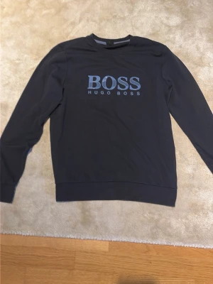 Mörkblå sweatshirt från Hugo Boss - Snygg mörkblå sweatshirt från Hugo Boss med stor logga i ljusblått framtill. Tröjan har rund halsringning, långa ärmar och ribbade muddar vid ärmslut och nederkant. Perfekt för en clean och stilren look. Sitter som M
