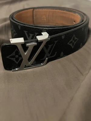 Svart  monogram skärp - Säljer ett svart skärp  med klassiskt monogrammönster och en stor L logga i silver som spänne. Skärpet är gjort i skinn och har en lyxig känsla som verkligen sticker ut.