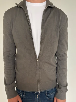 Grå ziptröja med hög krage - Snygg grå ziptröja med hög krage och ribbade muddar. Tröjan har dragkedja hela vägen och är tillverkad i ett mjukt, tunt material som passar perfekt till jeans eller över en t-shirt. Enkel och stilren design utan synligt märke.