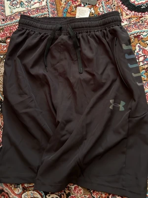 Svarta träningsshorts från Under Armour - Snygga svarta träningsshorts från Under Armour med elastisk midja och snörning. Shortsen har logga på vänster ben och diskreta detaljer längs sidan. Tillverkade i ett lätt och snabbtorkande syntetmaterial, perfekta för sport och aktivitet.
