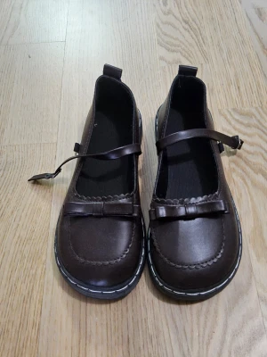 Bruna  skor med rosett - Mörkbruna skor i konstläder med rundad tå, dekorativ rosett framtill och justerbara remmar över foten. Skorna har kontrastsömmar längs sulan och en klassisk, söt design som passar till många olika stilar. Skulle säga den passar 36. Skriv om du vill ha flera bilder eller mer info om skorna