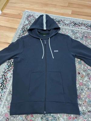 Mörkblå zip hoodie från BOSS - Mörkblå hoodie med dragkedja från BOSS. Huva med vita snören och vit rand på toppen, samt vita detaljer på muddarna. Fickor framtill och diskret BOSS-logga på bröstet. Perfekt för en avslappnad och stilren look.