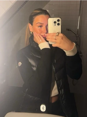 Svart dunväst från Moncler - Snygg svart ”dunväst” med ärmar från Moncler med klassisk logga på ärmen. Västen har quiltad framsida, hög krage och dragkedja. Perfekt för lager-på-lager och ger en stilren look. Materialet är glansigt och dunfyllt framtill, med mjuka svarta ärmar. Endast använd fåtal gånger nypris ligger upp mot 10 000kr, vid snabb affär kan jag gå ner lite 