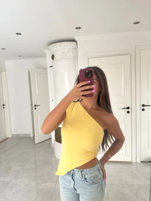 Gul oneshoulder topp med spänne - Snygg gul oneshoulder topp med croppad passform och dekorativt spänne i sidan. Perfekt för dig som vill sticka ut med en trendig och färgglad look. Tunn och mjuk känsla, passar till jeans eller kjol.