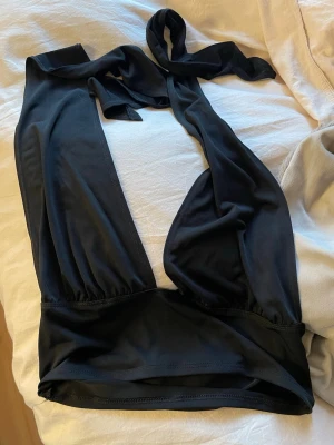 Svart omlottopp från NLY One - Säljer en svart omlottopp från NLY One i mjukt, tunt material. Toppen har breda axelband och knytband i nacken för en snygg halterneck-look. Perfekt att styla med höga jeans eller kjol för en trendig outfit.