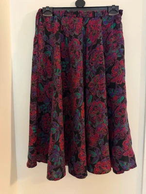 Blommig vintage  midikjol med färgglada detaljer - Säljer en  vintage midikjol med färgglatt blommönster i rött, lila och grönt. Kjolen har en ledig passform och resår i midjan för extra komfort. 