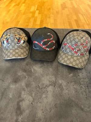 Gucci kepsar med tryck - 500 KRONOR STYCK! 1500 FÖR ALLA! Tre kepsar från Gucci med klassiskt GG-mönster. Två kepsar har ormtryck i rött, vitt och svart, och en har tigertryck framtill. Alla har svart mesh baktill och böjd skärm. Perfekt för dig som vill sticka ut med lyxig streetstyle.
