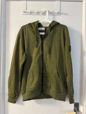 Olivgrön hoodie från Stone Island - Snygg olivgrön hoodie från Stone Island med dragkedja framtill, två fickor och klassisk logotyp på ärmen. Tillverkad i mjukt bomullsmaterial och har ribbade muddar samt justerbar huva med snören. Perfekt för en avslappnad streetwear-look.