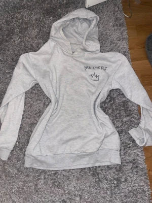 Ljusgrå hoodie med tryck Ma Chérie - Säljer en ljusgrå hoodie med huva och trycket 'Ma Chérie' samt en krona både fram och bak. Hoodien har ribbade muddar vid ärmslut och nederkant. Mjuk och skön, perfekt för chill dagar. Materialet känns som bomullsblandning.