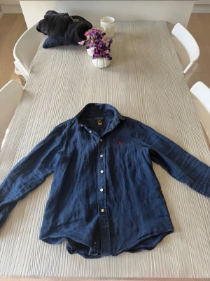Mörkblå linneskjorta från Polo Ralph Lauren - Snygg mörkblå skjorta i linne från Polo Ralph Lauren med klassisk krage och långa ärmar. Skjortan har vita knappar framtill och den ikoniska röda broderade loggan på bröstet. Perfekt för en avslappnad men stilren look.