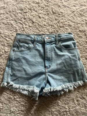 Ljusblå denimshorts med fransar - Säljer ett par ljusblå denimshorts från Wrangler med hög midja och råa fransade benslut. Klassisk femficksmodell med bälteshällor och knappgylf. Perfekta för varma dagar och en avslappnad stil.