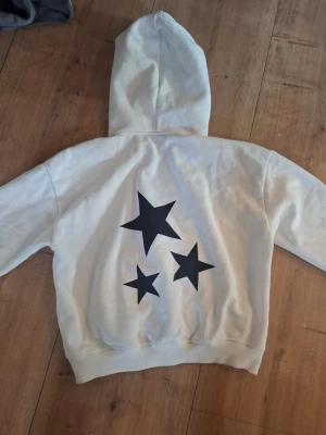Vit hoodie med svarta stjärnor XS - Säljer en vit hoodie i storlek XS med tre stora svarta stjärnor tryckta på ryggen. Hoodien har klassisk huva och ribbade muddar. Perfekt för dig som gillar enkel men cool design. Materialet är mjukt och skönt, troligen bomull eller bomullsblandning.