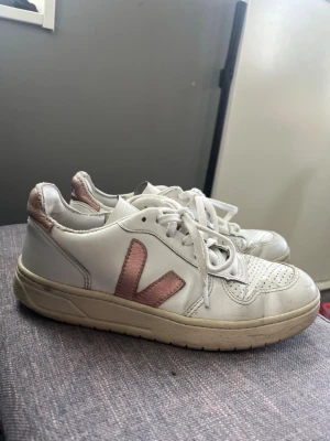 Vita Veja sneakers med rosa detaljer - Säljer ett par klassiska vita Veja sneakers med rosa metallic-detaljer på hälen och V-loggan på sidan. Skorna har perforerad tå, vita skosnören och en beige sula. Tillverkade i läder och passar perfekt till en trendig vardagslook.
