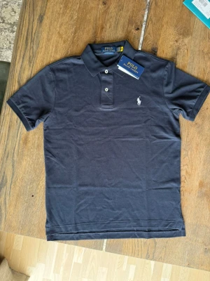 Mörkblå pikétröja från Polo Ralph Lauren - Klassisk mörkblå pikétröja från Polo Ralph Lauren med vit broderad logga på bröstet. Tröjan har krage, knappar framtill och korta ärmar. Tillverkad i mjuk bomull som känns skön mot huden.