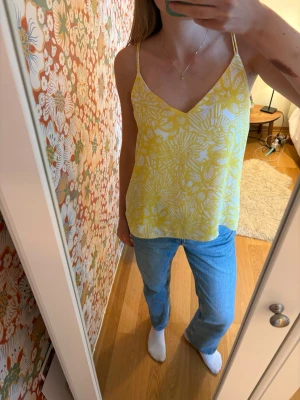 Gul blommig linnetopp med smala band - Snygg gul linnetopp med vitt blommigt mönster och smala axelband som korsas i ryggen. Toppen har v-ringning och är tillverkad i ett lätt material, perfekt för varma dagar. Passformen är lös och luftig.