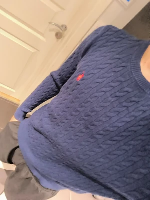 Mörkblå kabelstickad tröja Polo Ralph Lauren - Klassisk mörkblå kabelstickad tröja från Polo Ralph Lauren med rund halsringning och den ikoniska röda logotypen broderad på bröstet. Tröjan har långa ärmar och ribbade muddar vid ärmslut och nederkant. Perfekt för en stilren och tidlös look.