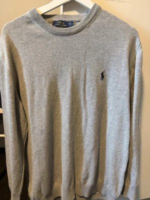 Grå stickad tröja från Polo Ralph Lauren - Snygg grå stickad tröja från Polo Ralph Lauren med rund halsringning och klassisk broderad logga på bröstet. Tröjan har ribbade muddar vid ärmslut och nederkant, perfekt för en stilren look. Materialet är mjukt och bekvämt.