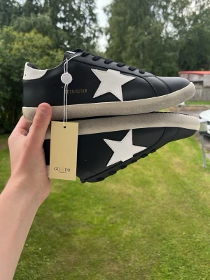 Svarta Golden Goose sneakers med vit stjärna ÄKTA - Helt nya och HELT OANVÄNDA!🔥Helt oanvända med taggar kvar.‼️ Säljs till ett lågt pris för snabb affär.‼️Svarta sneakers från Golden Goose med vit stjärndetalj på sidan och vit häl. Skorna har svarta skosnören och en beige sula. Kvitto följs inte med när man köper, visas bara på bilden för bevis. Tillverkade i läder och levereras med originalkartong och tillbehör. Perfekta för en trendig och avslappnad stil.