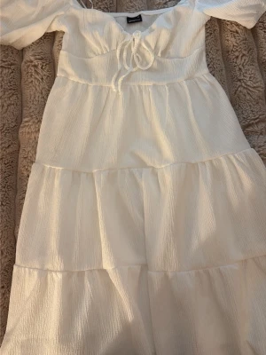 Vit omlottklänning från Gina Tricot - Söt vit klänning från Gina Tricot med puffärmar, omlottdetalj och knyt framtill. Klänningen har en luftig, volangig kjol och är tillverkad i ett lätt, strukturerat tyg som passar perfekt till varma dagar.