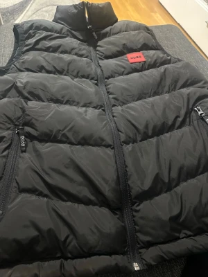 Svart dunväst från Hugo Boss - Snygg svart dunväst från Hugo Boss med hög krage och dragkedja framtill. Västen har två sidofickor med dragkedja och en röd patch med HUGO på bröstet. Perfekt för lager-på-lager och riktigt trendig look.