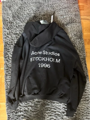 Svart hoodie Acne Studios Stockholm - Säljer en svart hoodie från Acne Studios med tryck på ryggen där det står 'Acne Studios STOCKHOLM 1996'. Hoodien har en klassisk huva och ribbade muddar vid ärmslut och nederkant. Perfekt för dig som gillar stilrena och trendiga plagg. Aldrig använd, bara testad. Säljer då den är för stor. Pris kan diskuteras.