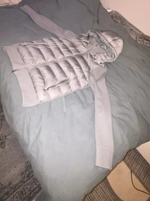 Ljusgrå quiltad cardigan med huva - Snygg ljusgrå cardiganjacka med quiltad kropp och stickade ärmar. Jackan har dragkedja framtill, två fickor med dragkedja och en mysig huva. Perfekt för lager på lager och chill stil. Materialmix av vadderad polyester och stickat tyg. Riktigt skön och varm cardigan.