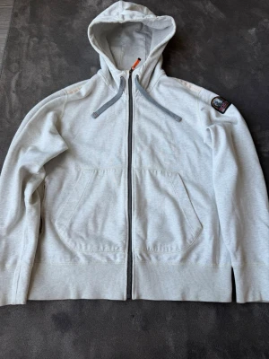Ljusgrå hoodie från Parajumpers - Ljusgrå hoodie från Parajumpers med dragkedja, huva och gråa dragsnören. Fleecefodrad insida för extra komfort och värme. Två stora fickor framtill och en patch på ena ärmen. Orange detaljer vid dragkedjan ger en cool kontrast.
