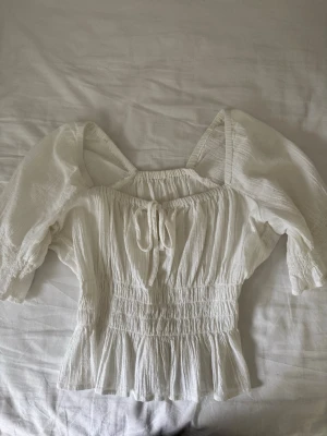 Vit blus med puffärm - Söt vit blus med puffiga ärmar och smockad midja. Blusen har en liten volangkant nedtill och knytning fram. Jag har tidigare sytt in ryggen. På sista bilden syns märken som kan synas. Från H&M, vet tyvärr inte storlek men passar ungefär S. 