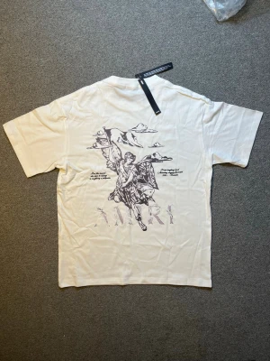 Vit t-shirt med ängeltryck från Amiri - Cool vit t-shirt från Amiri med stort grafiskt tryck av en ängel och moln på ryggen. T-shirten har korta ärmar och en avslappnad passform. Perfekt för dig som gillar streetwear och unika prints.