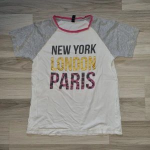 Vit och grå t-shirt med text - En vit och grå t-shirt med texten 'NEW YORK', 'LONDON' och 'PARIS' i svart, gult och rosa. T-shirten har korta ärmar och en rosa kant runt halsen. stl 146/152