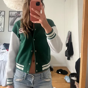 Grön och vit bomberjacka - En grön och vit colleagejacka med knappar framtill. Jackan har långa ärmar och ribbade muddar vid ärmslut och nederkant. 