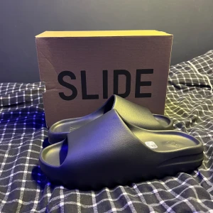 Yeezy Slide Dark Onyx - Säljer ett par yeezy slides i colorwayen Dark Onyx. De är i storlek 47 men är små i storleken så de passar 46 också. De är helt nya deadstock och aldrig använda, tog de bara ut ur lådan för att ta bilderna. Pris kan diskuteras.