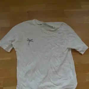 En vit t- shirt med en liten gullig Palm vid bröstet. Tröjan ser lite skrynklig ut på bilden men lämnas såklart strycken 