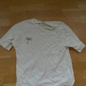 Vit t-shirt med litet tryck  - En vit t- shirt med en liten gullig Palm vid bröstet. Tröjan ser lite skrynklig ut på bilden men lämnas såklart strycken 