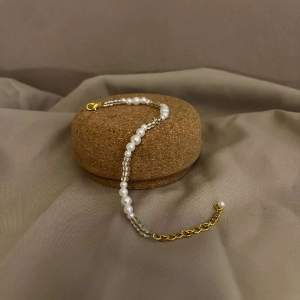 Beskrivning: Detta armband heter Thimea och är ett armband med olika storlekar av vita pärlor och beiga pärlor med lås och kedja i guldplätering. Namn: Thimea Längd: Hela 22,5 cm Pärlor 15 cm 
