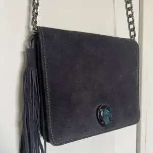 En mörkblå handväska i mocka från Caroline Svedbom. Med en mörkblå Sten och grå dragkedja. En toffs med fransar på ena sidan.  Fint skick då den endast är använd fåtal gånger. 