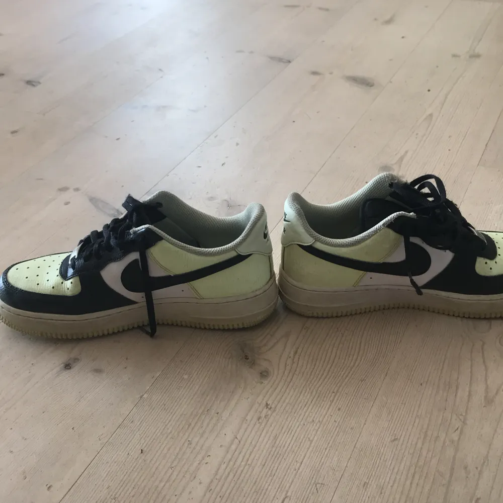 Ett par Nike Air Force 1 sneakers i svart, vit  och ljusgul färg. Skorna har snörning och en perforerad tåbox för ventilation. De är i storlek EUR 40, 25.5 cm. Skor.