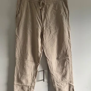 Beige linnebyxor - Ett par beige linnebyxor i storlek M. Byxorna har en elastisk midja med snörning för justerbar passform. De är tillverkade av en blandning av linne och bomull, vilket gör dem bekväma och andningsbara.
