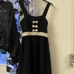 Svart klänning med vita detaljer - En elegant svart klänning med vita detaljer. Klänningen har breda axelband och tre dekorativa rosetter på bröstet. Den har en vit midjeband som framhäver midjan.